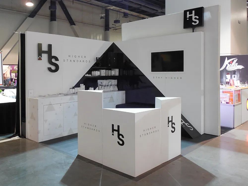 Higher Standards 10x20 inline trade show booth with white angular walls, black triangle graphic, product shelving and reception counter