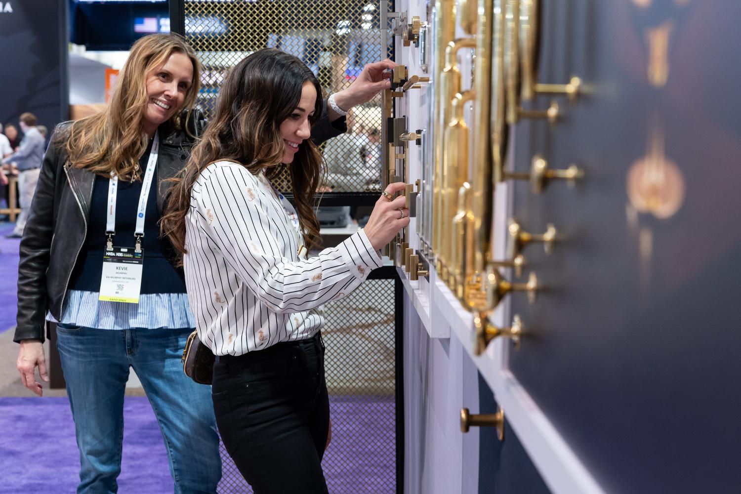 Two KBIS attendees testing brass cabinet hardware samples mounted on a display wall