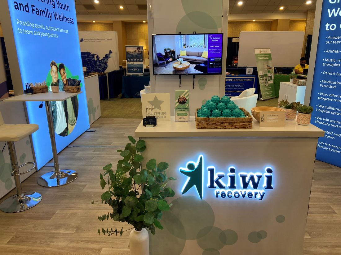Kiwi Recovery 10x10 inline trade show booth with backlit light blue graphics and a branded counter