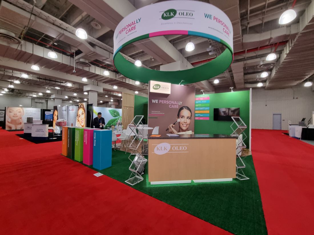 KLK Oleo 20x20 island trade show exhibit with a circular hanging sign, color-coded product pedestals and a seating area