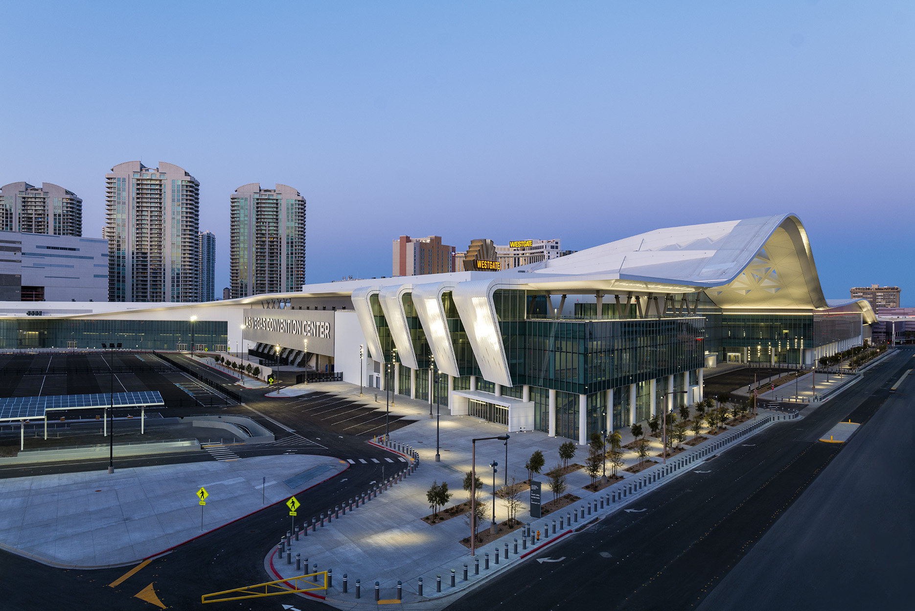 The Las Vegas Convention Center West Hall lit at dusk, with the exhibit halls, entrance plaza and surrounding Paradise Road properties visible