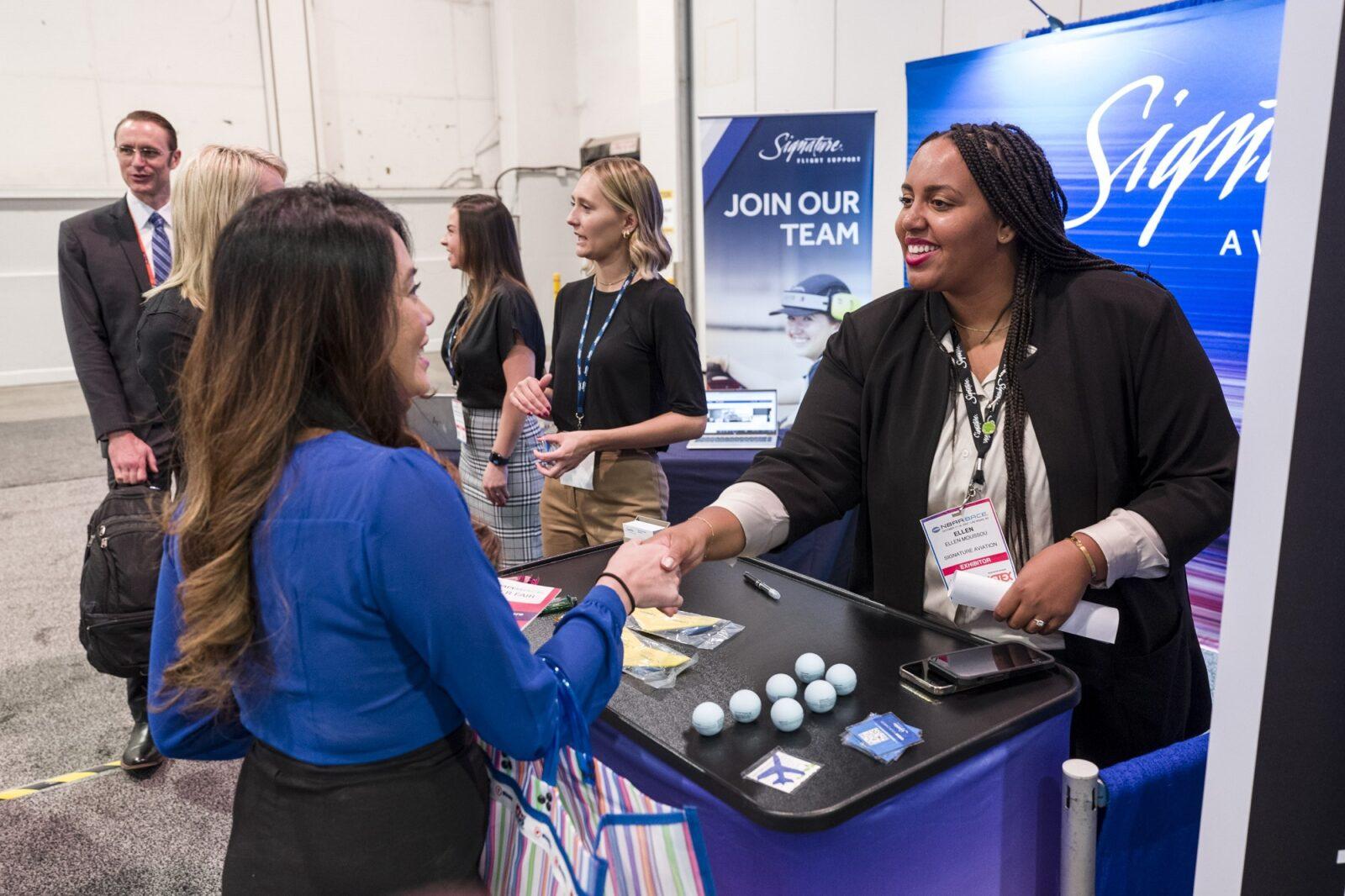An exhibitor greets a visitor with a handshake at a staffed counter on a trade show floor.