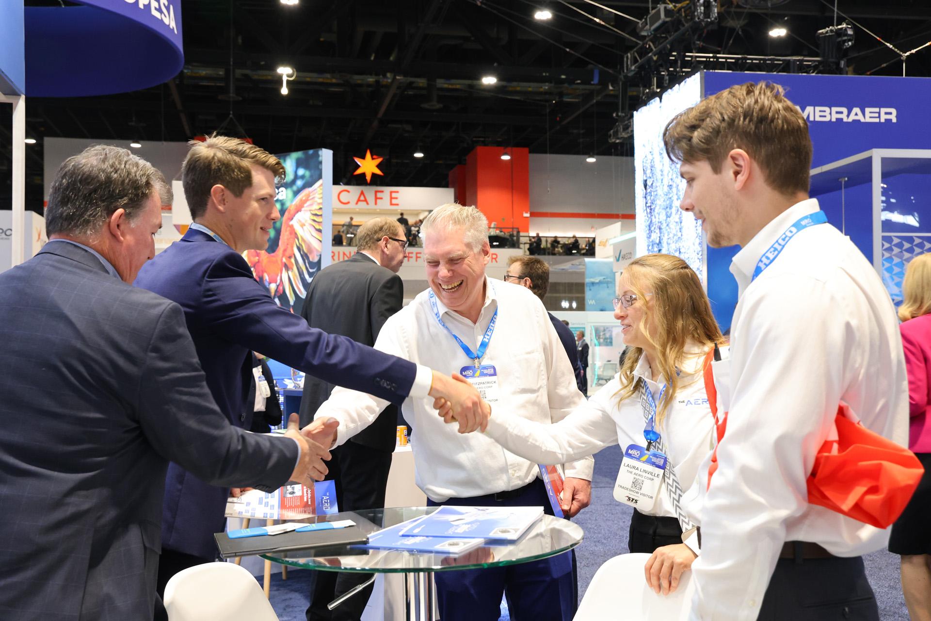 Exhibitors and visitors shake hands across a meeting table at an MRO Americas exhibit stand.