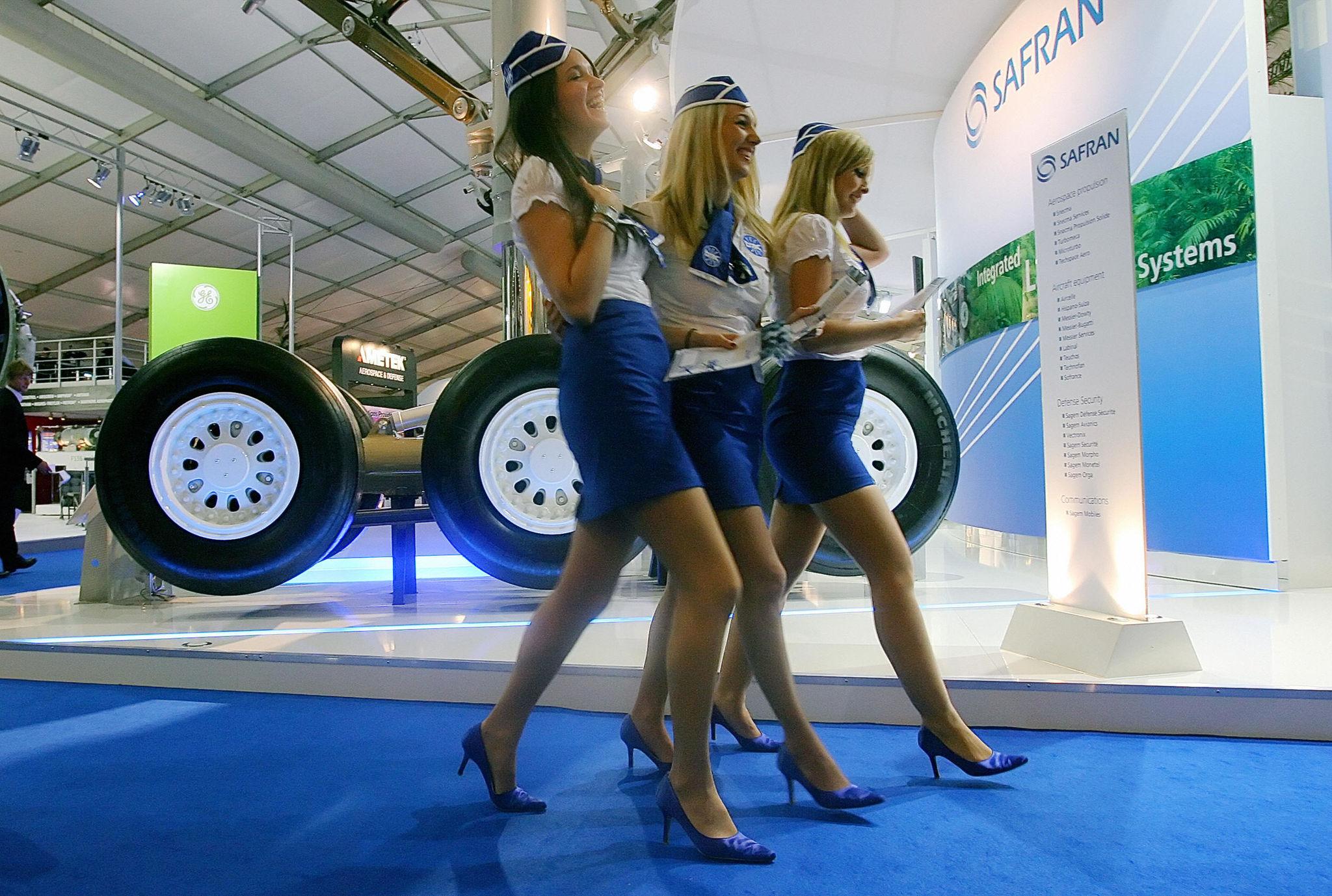 Three uniformed exhibit hosts walk past an aerospace supplier stand with aircraft landing gear on display.