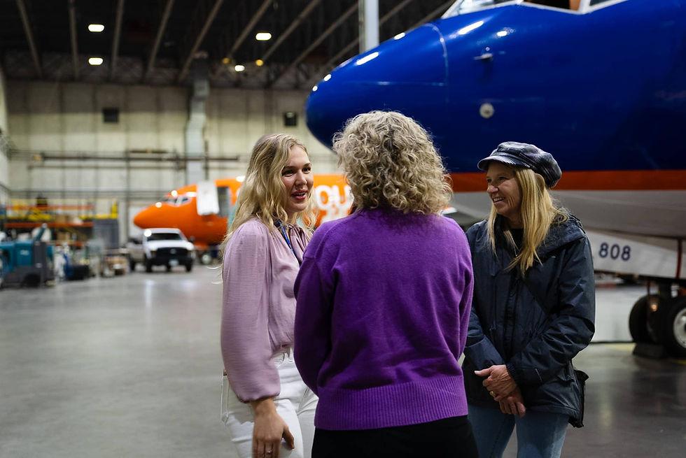 Three aviation professionals talk together inside a maintenance hangar with aircraft parked behind them.
