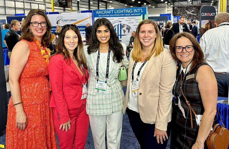 Five aviation professionals stand together in an exhibition aisle with exhibitor stands and signage behind them.