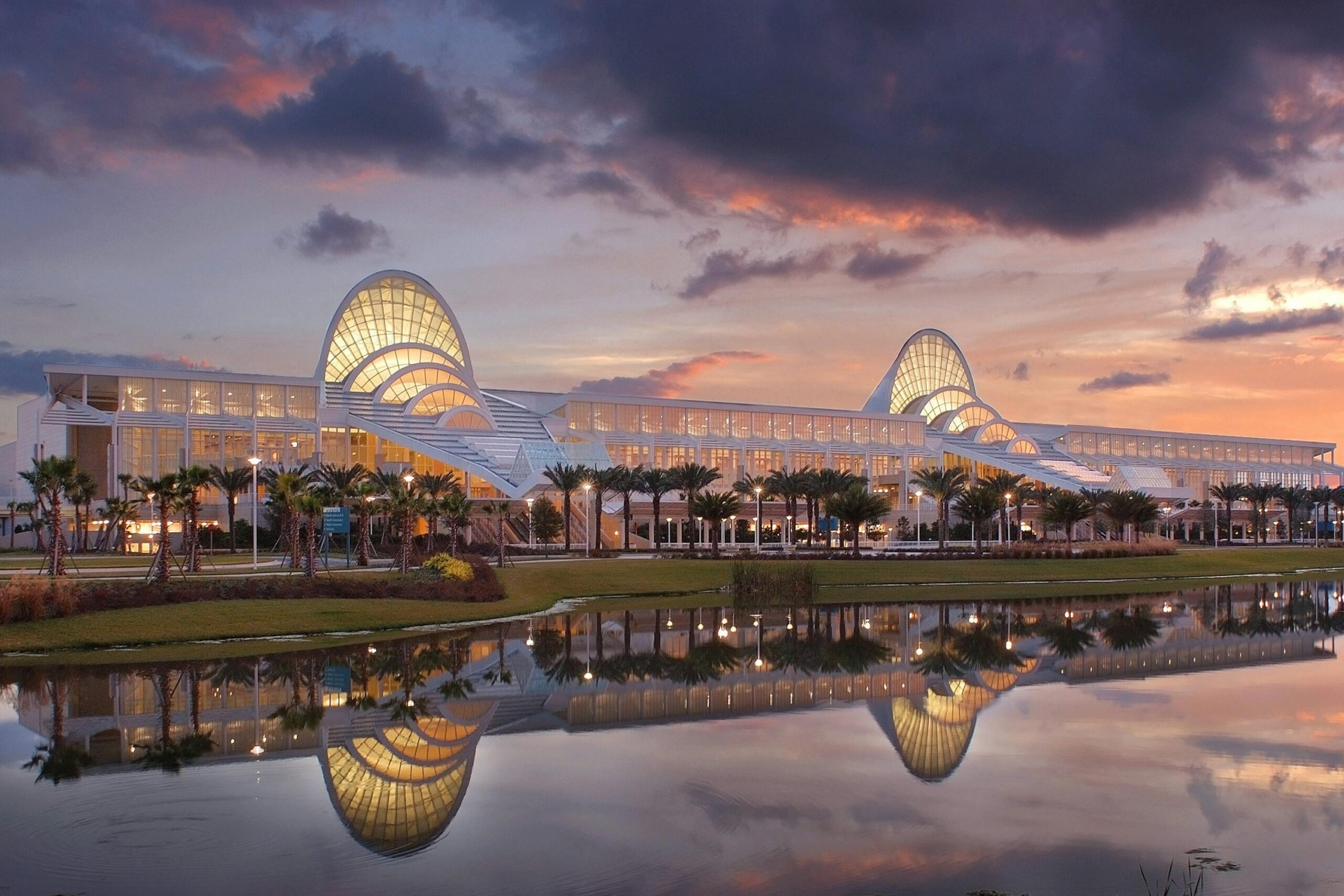 The Orange County Convention Center in Orlando at dusk, its curved glass entrances lit from within and reflected in the pond in front of the building.
