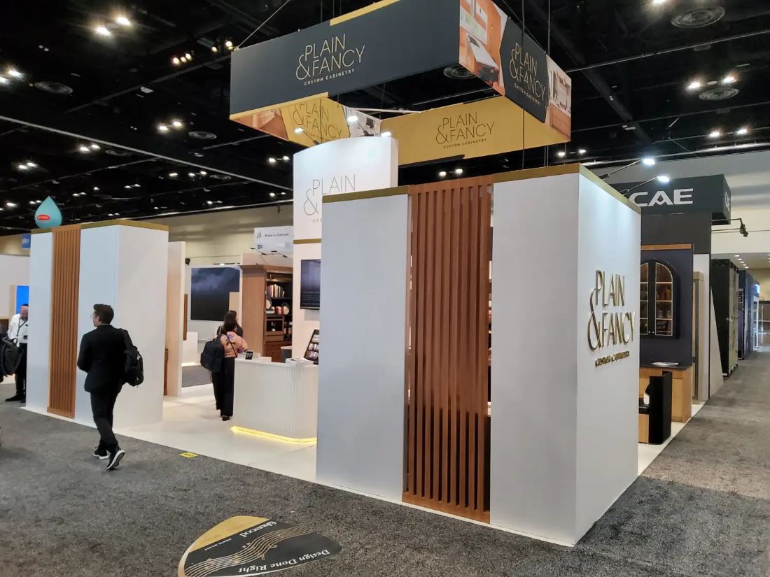 Plain and Fancy 20x30 island trade show exhibit with white towers, wood slat accents, hanging fabric signs and kitchen vignettes