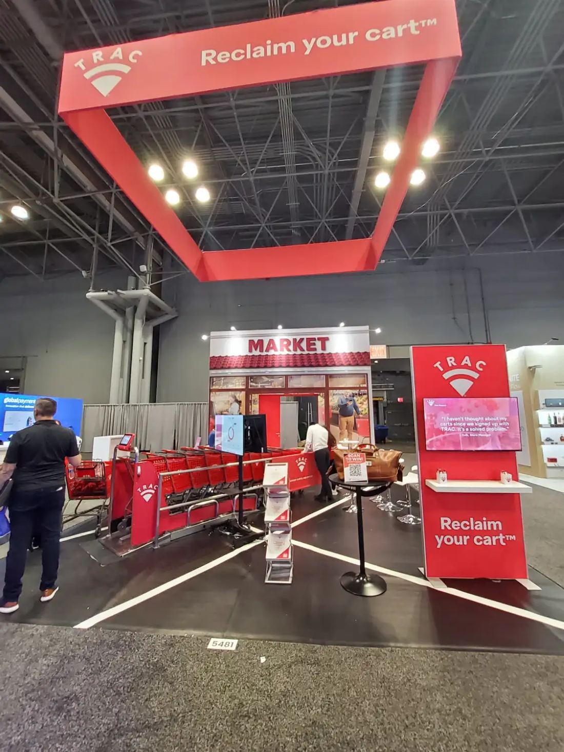 TRAC 20x20 island trade show booth with a red hanging frame sign, storefront facade and working shopping cart demo