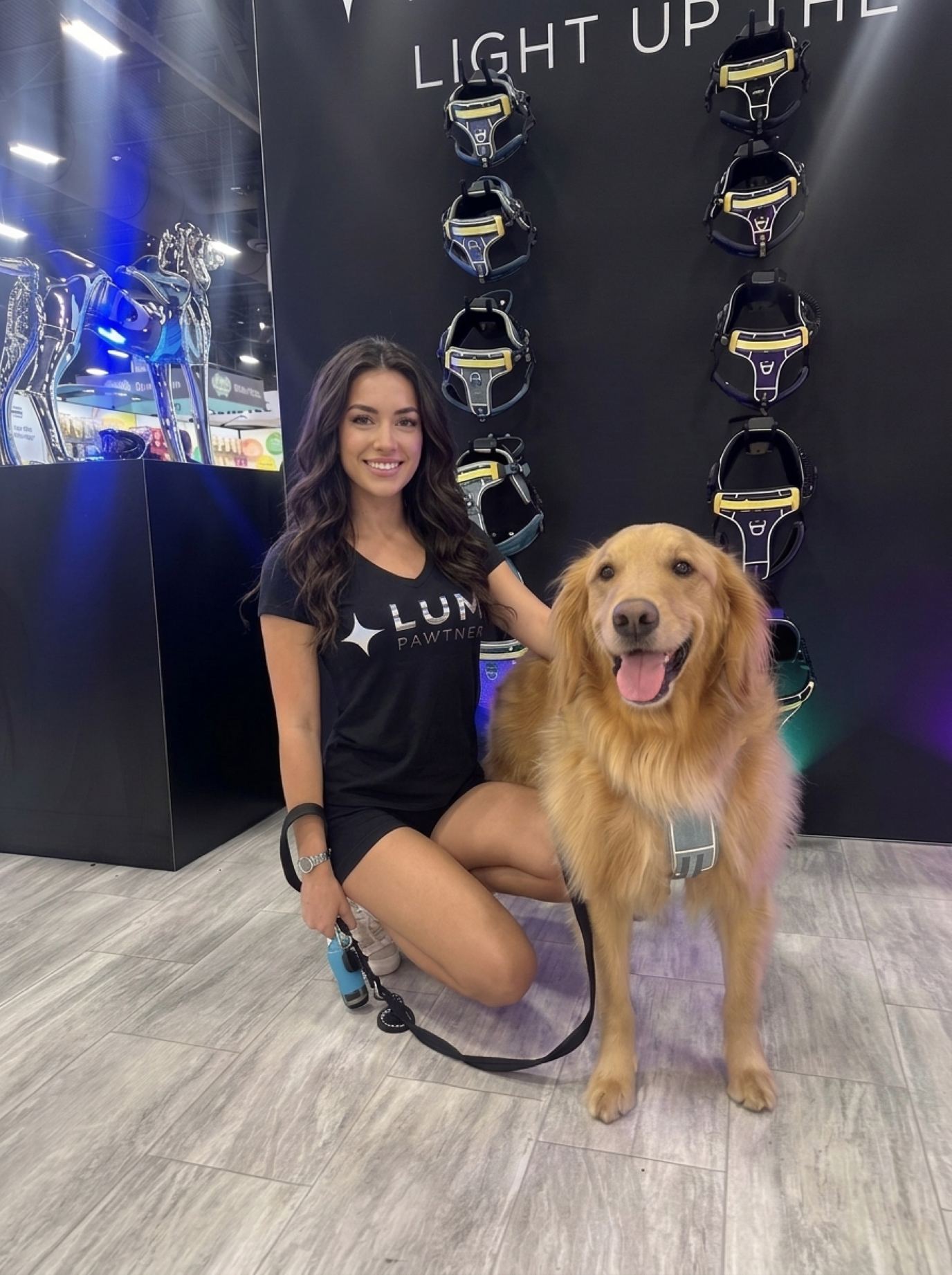 Brand ambassador in a black logo tee kneeling beside a golden retriever wearing a harness in front of a wall of dog harnesses