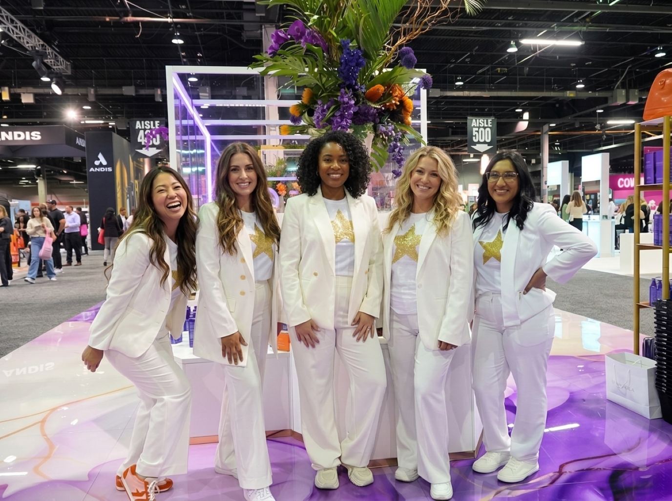 Five promotional models in matching white pantsuits over star-print tees, smiling together on a glossy floor at a beauty booth