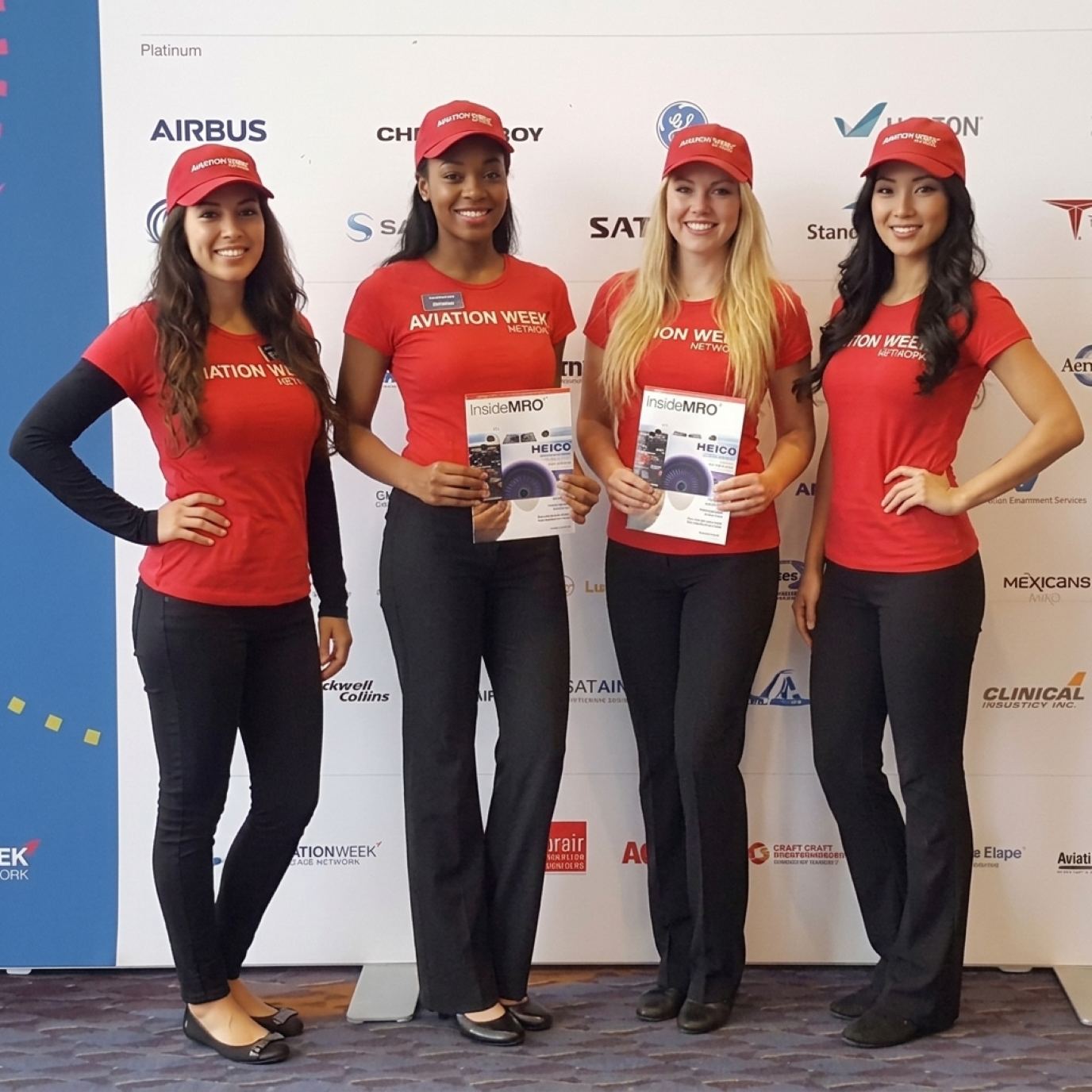 Four brand ambassadors in red caps, fitted red logo tees and black trousers holding magazines in front of a sponsor wall, a bold branded uniform idea for SHOT Show trade show models
