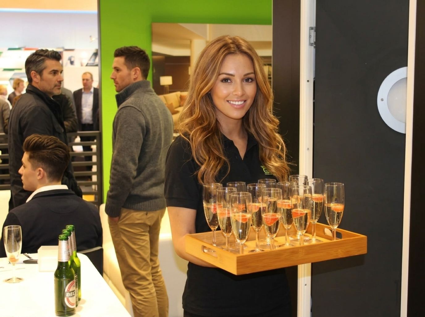 Hostess in a black logo polo carrying a bamboo tray of sparkling flutes topped with strawberries through a busy exhibit lounge, the tray service we bring to Winter Fancy Food Show booths