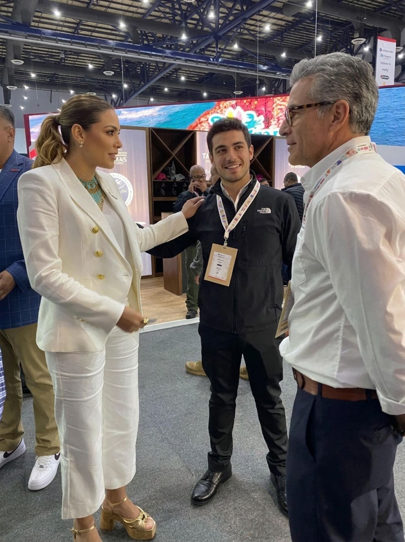 Hostess in a cream double-breasted blazer with gold buttons and wide cream trousers greeting two guests in front of a booth with an LED header