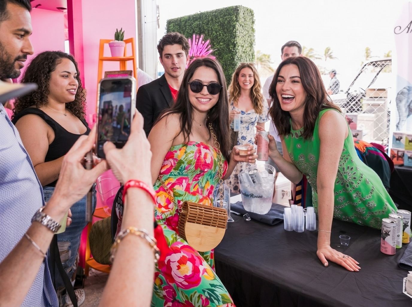 Hostess in a green dress leaning over a sampling counter while a guest in a floral dress poses for a phone photo, the kind of photo moment Licensing Expo booth staff run