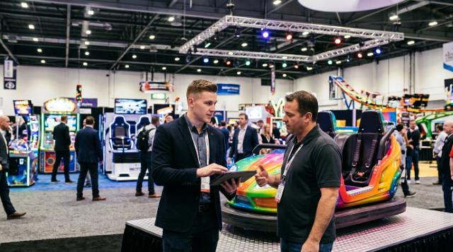 Booth rep in a dark suit showing a tablet to a visitor in a black polo next to a colorful ride vehicle at an amusement industry expo