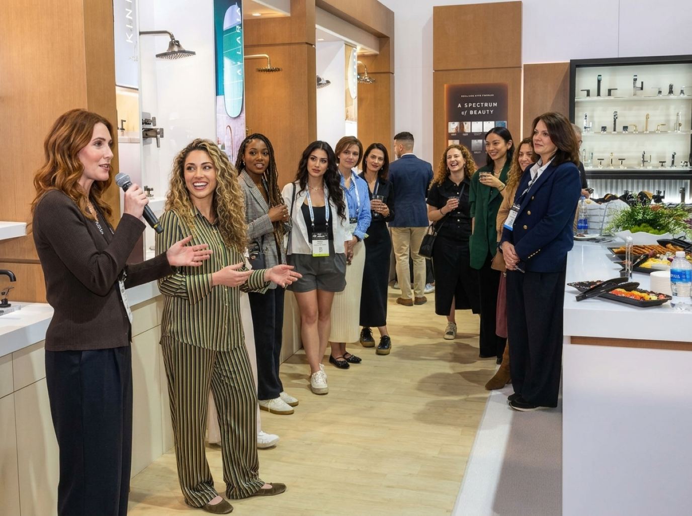 Purple Exhibits product demonstrators: a presenter in a brown ribbed cardigan with a handheld mic and a co-presenter in a striped satin set walk guests through a bath fixtures showroom booth