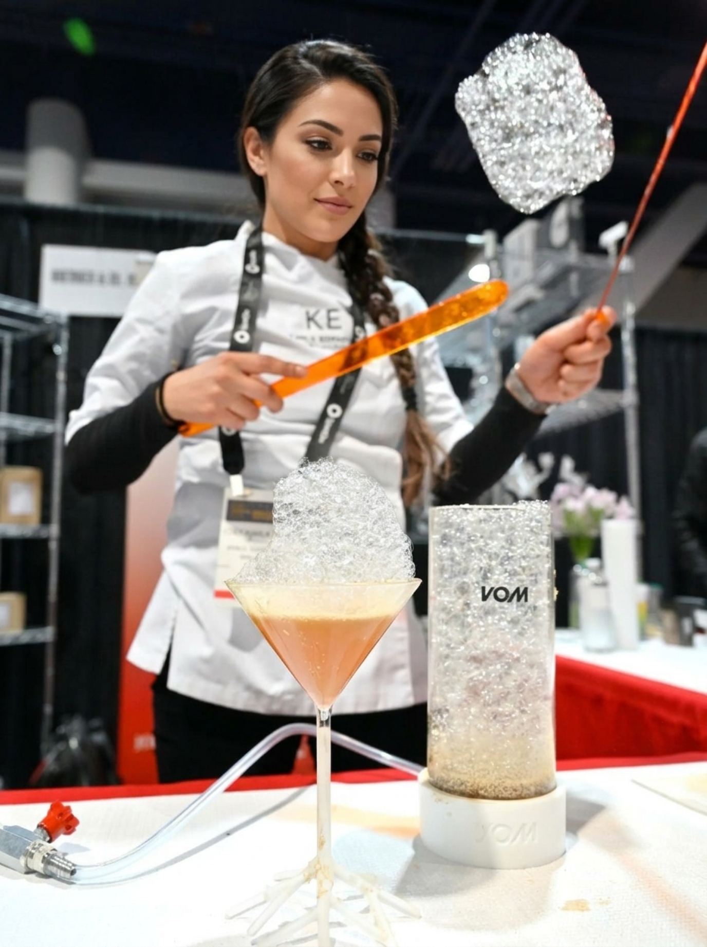 Demonstrator in a white chef jacket and lanyard lifting a cloud of foam above a cocktail glass at a beverage counter, product demonstrators for trade shows in a crisp culinary look