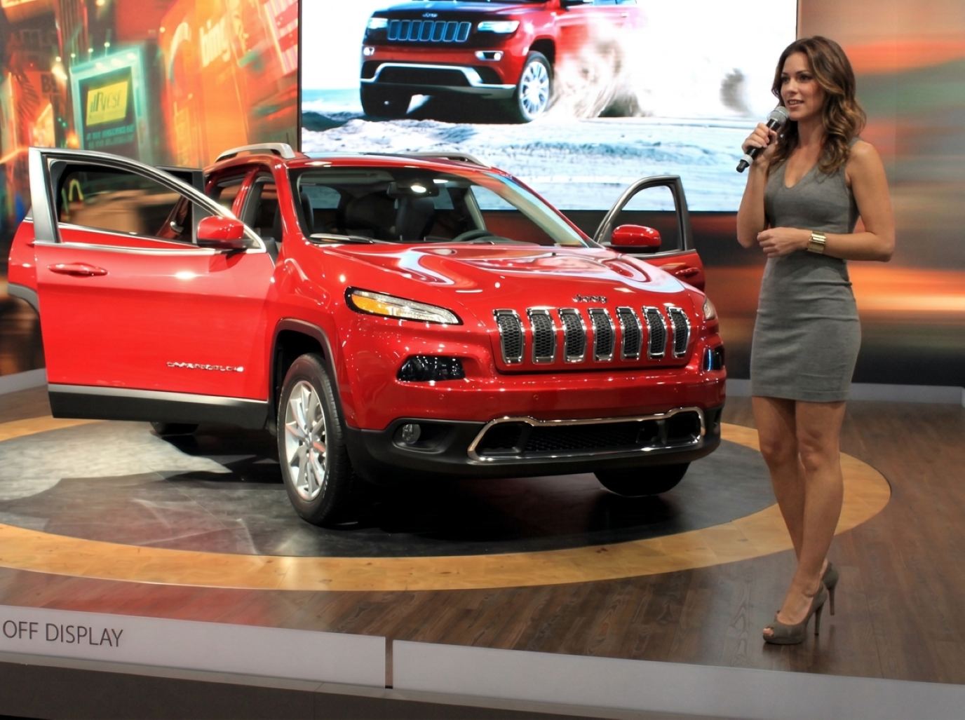 Product presenter in a gray dress speaking into a microphone beside a red SUV on a turntable stage