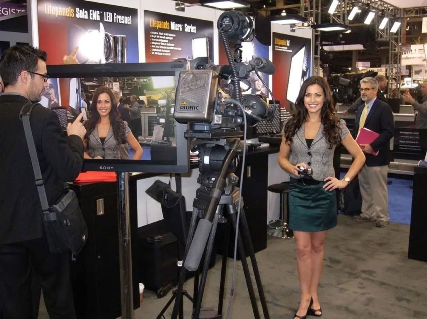 Product presenter in a gray tailored jacket and teal skirt holding a handheld mic beside a broadcast camera at a lighting and video booth, the presenter style we send to NAMM
