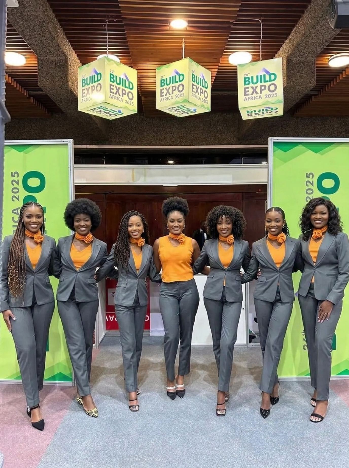 Seven hostesses in matching charcoal suits and orange scarves at the entrance of a building-products expo