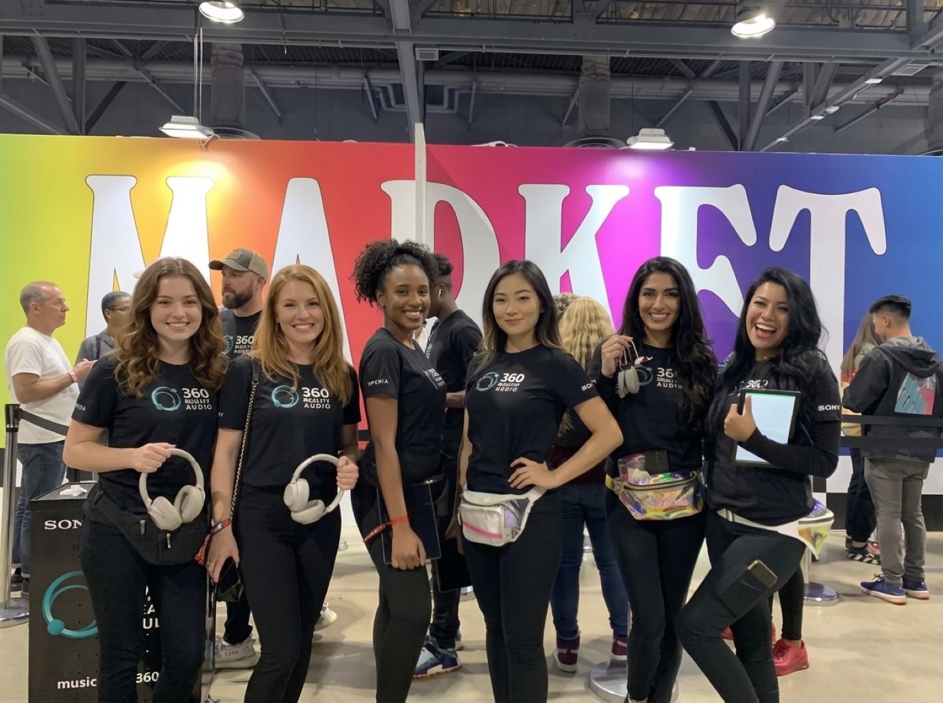 Six brand ambassadors in matching black tees and leggings holding headphones and a tablet in front of a bright wall, the kind of Black Hat booth staff crew Purple Exhibits books