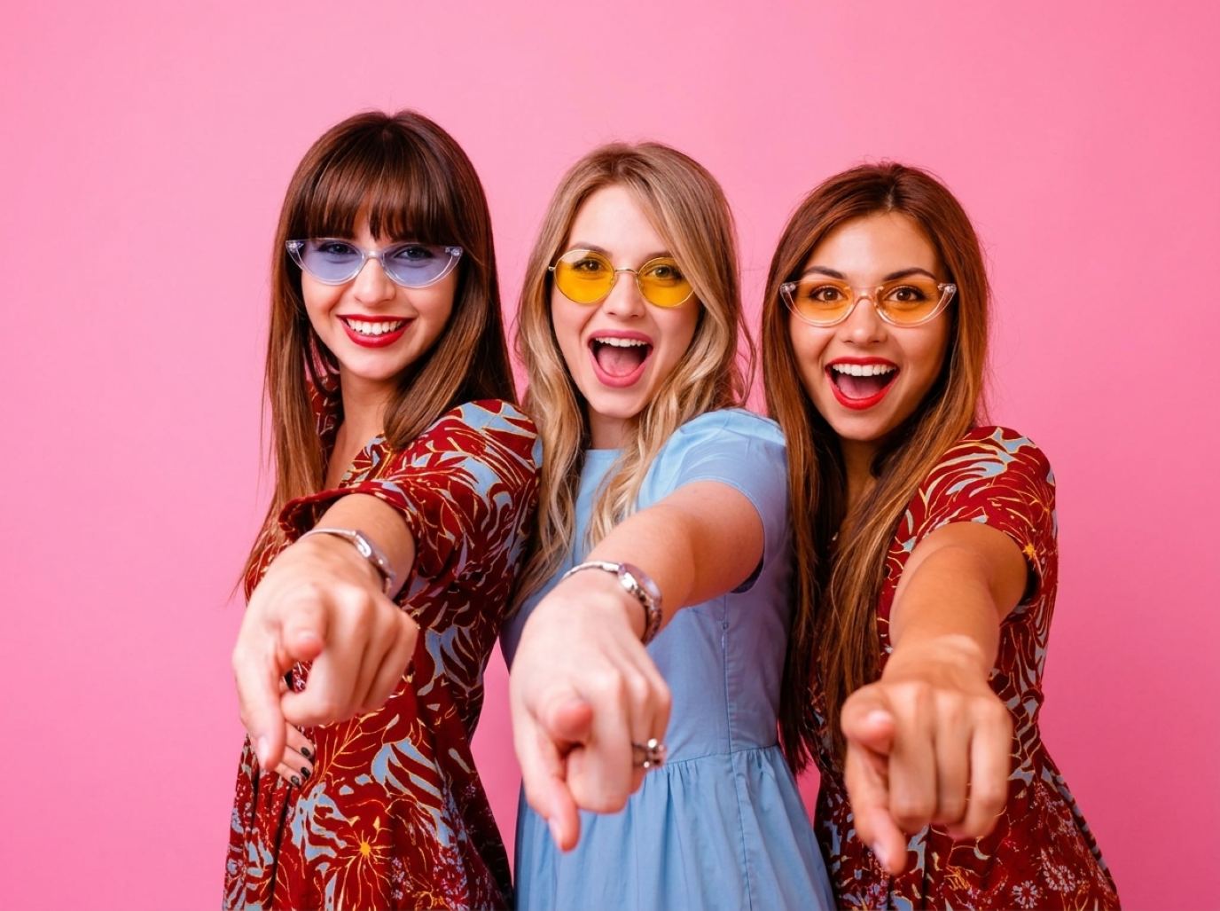Three Licensing Expo trade show models in bright printed dresses and colored sunglasses pointing toward the camera against a pink backdrop