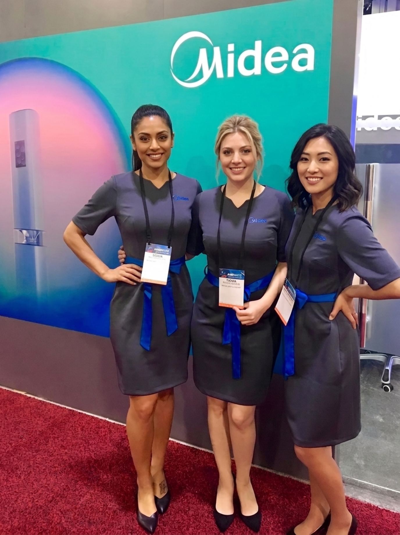 Three PACK EXPO booth staff in matching charcoal shift dresses with royal blue sash belts and lanyards, smiling on a red carpet in front of a lit exhibit wall