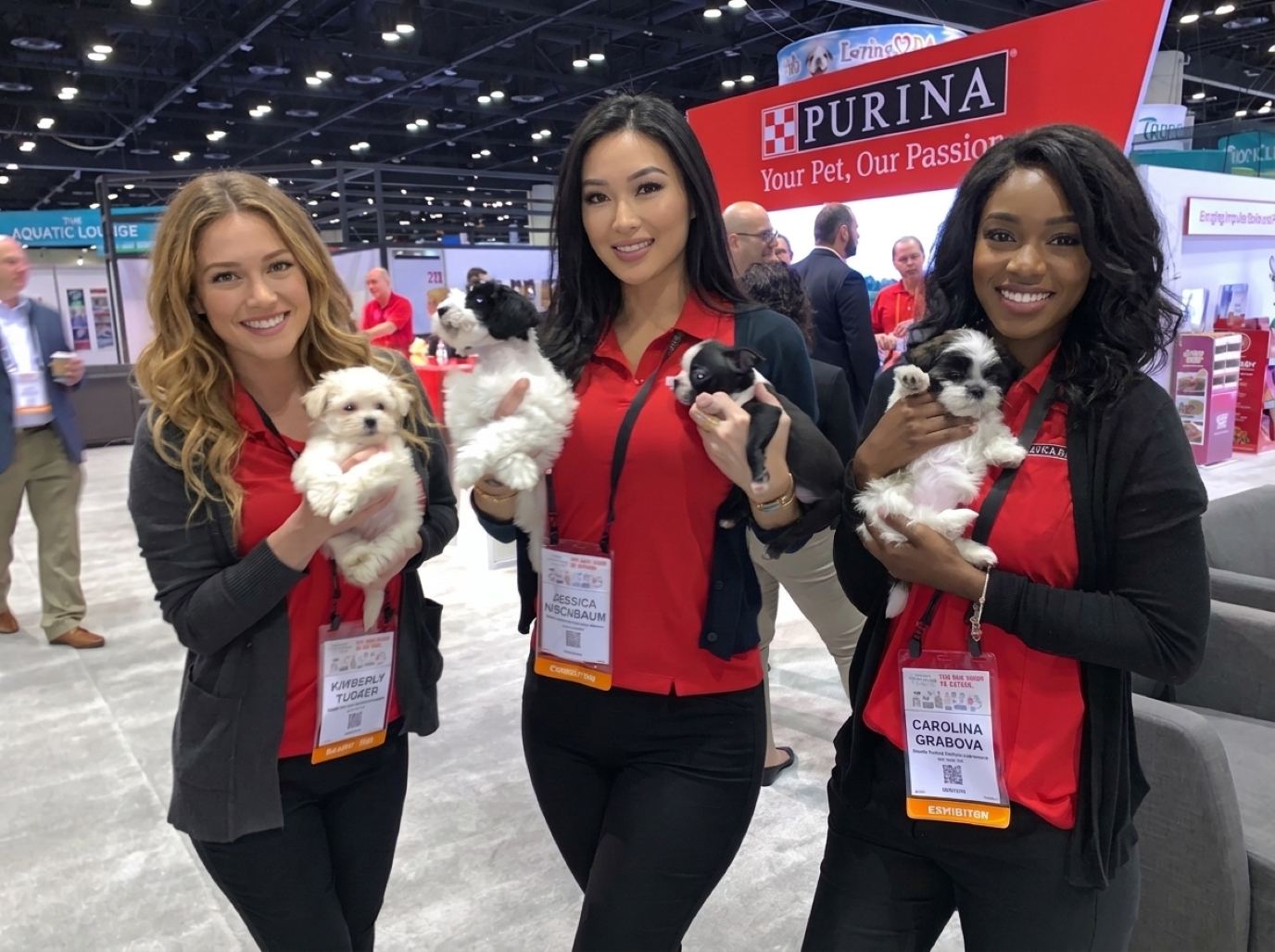 Three Global Pet Expo booth staff in red polos and charcoal cardigans smiling as they hold small puppies for a photo at a pet food booth