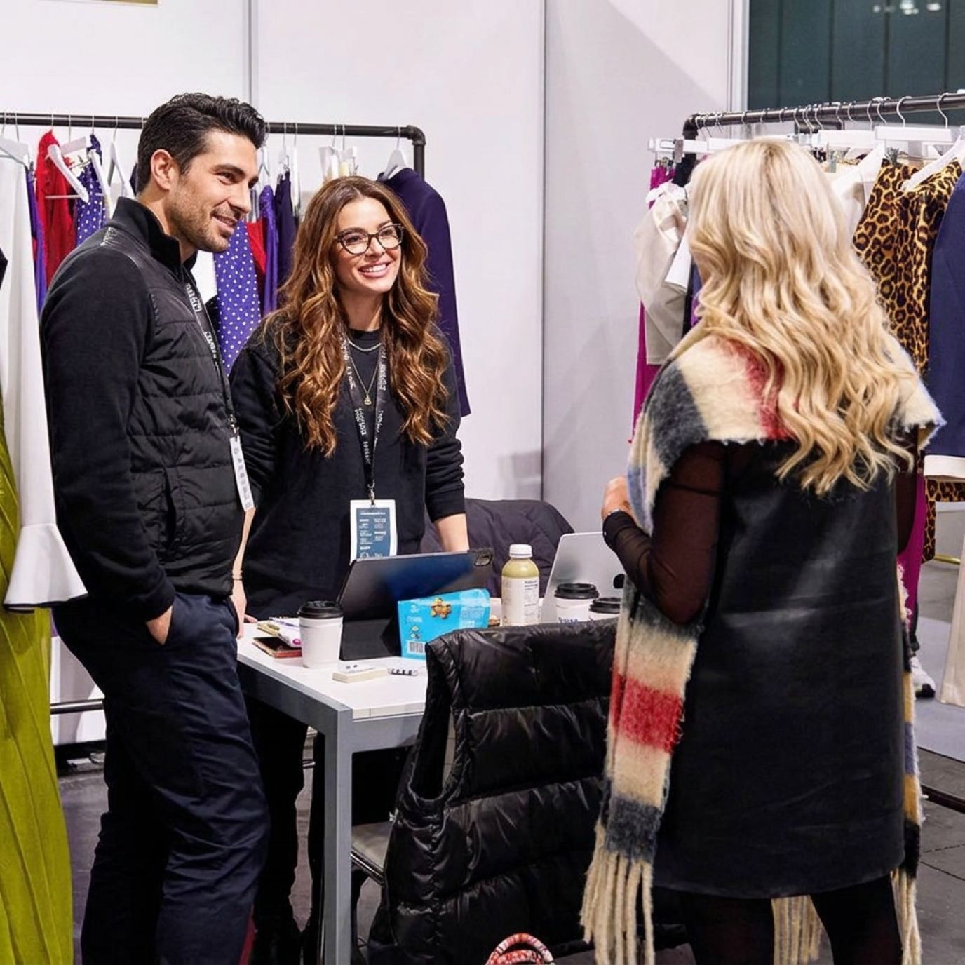 Two booth hosts in black with show lanyards chatting with a buyer between clothing racks at a fashion booth
