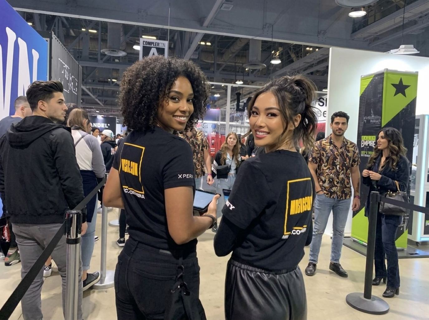 Two brand ambassadors in matching black logo tees holding tablets beside a roped queue line in a busy exhibit hall