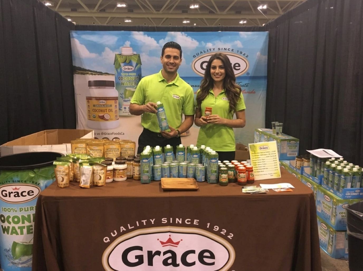 Two brand reps in lime green logo polos holding up a carton of coconut water and a jar of sauce behind a stocked food brand counter, the product-in-hand style of our Winter Fancy Food Show trade show models