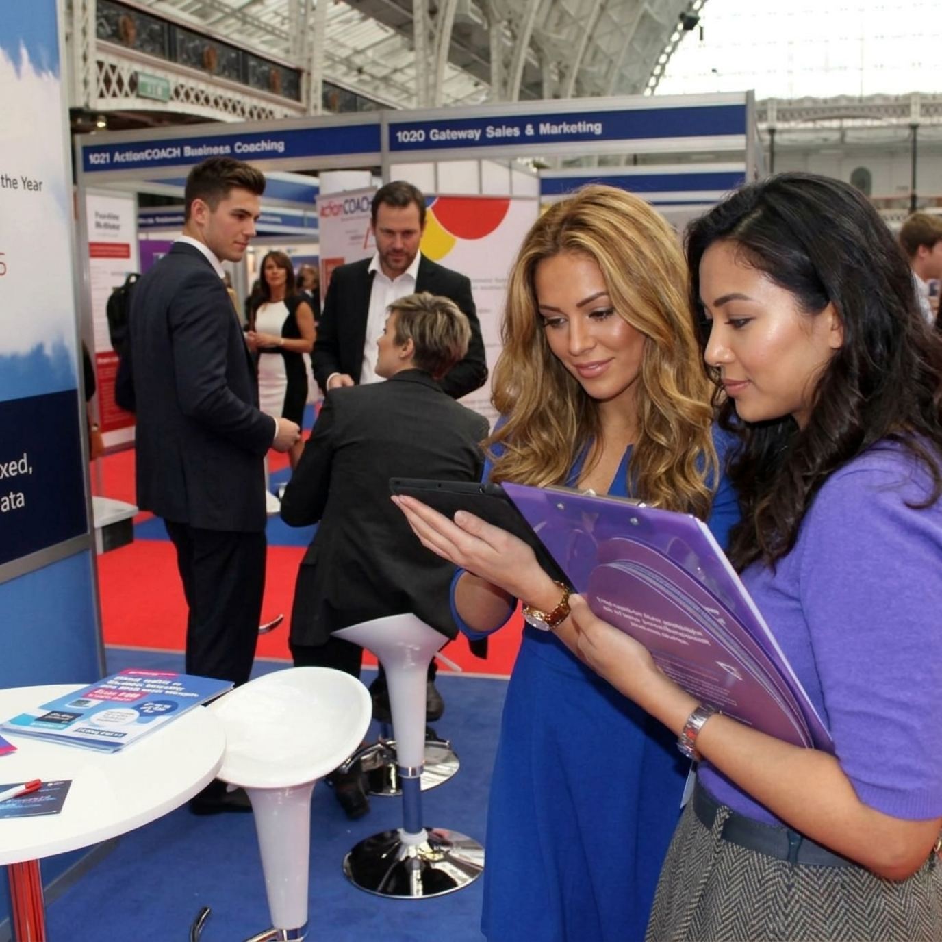 Two hostesses in matching royal blue checking a tablet and brochures at a business expo booth