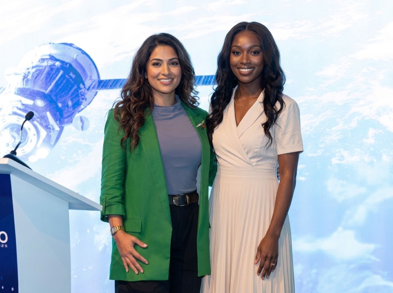 AWS re:Invent trade show models from Purple Exhibits: a stage host in an emerald blazer and a host in a cream pleated dress beside a podium in front of a space-themed screen