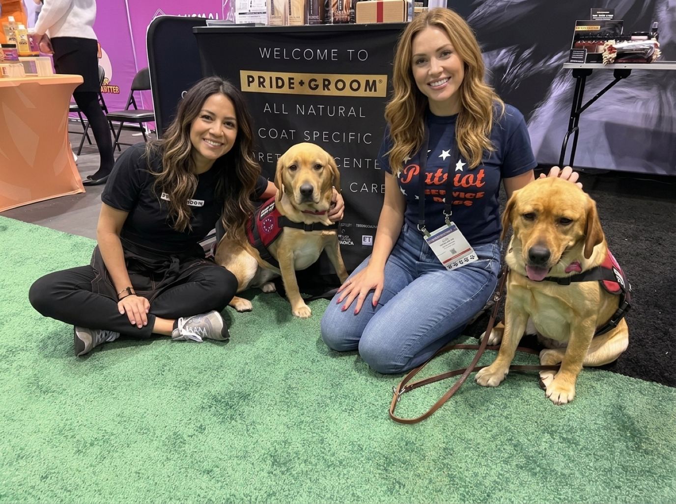 Two SuperZoo booth staff in dark tees kneeling on green turf beside two calm yellow Labradors in working vests at a natural grooming products booth