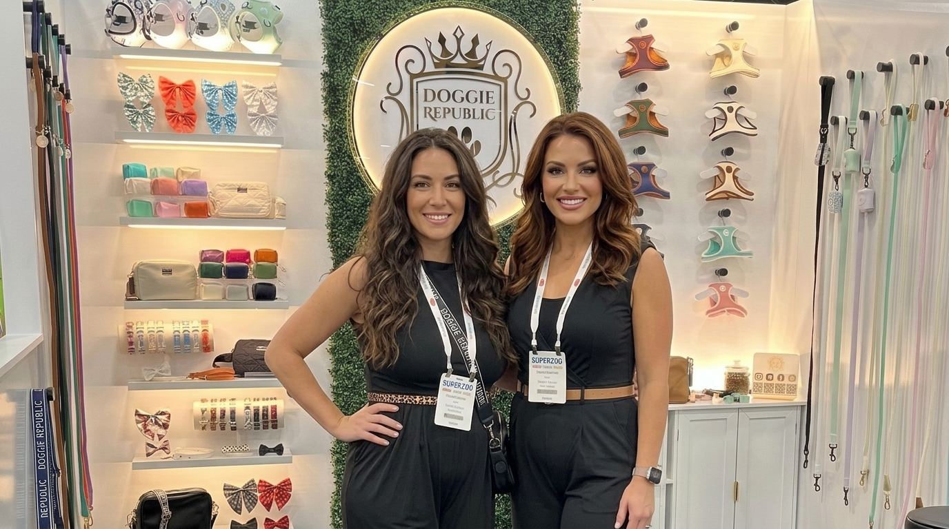 Two hosts in black jumpsuits with leopard and tan belts standing at a pet accessories booth with lit shelves of harnesses, bows and leashes, the polished look of Global Pet Expo trade show models